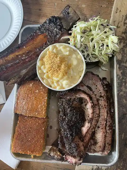A metal tray holds a large, charred rack of ribs, sliced smoked brisket, two pieces of cornbread, a bowl of macaroni and…