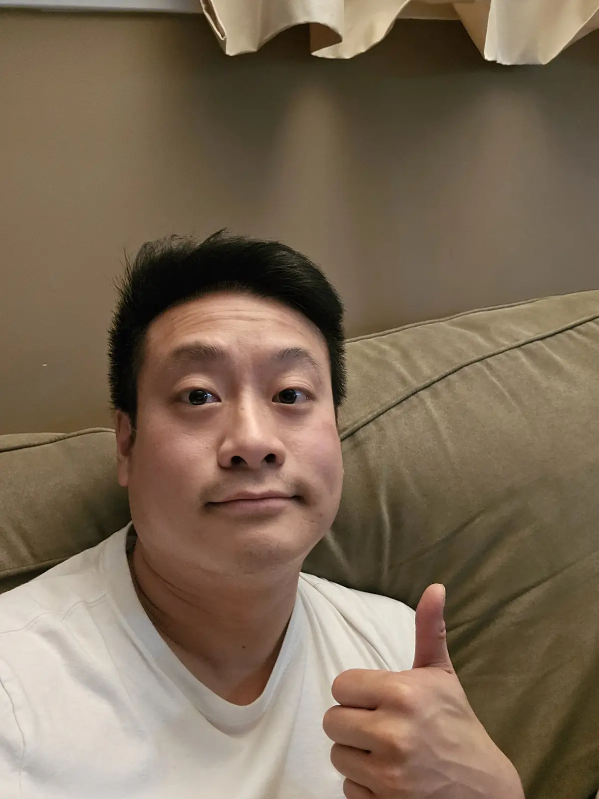 A man with short dark hair sits on a beige couch, wearing a white t-shirt, and gives a thumbs-up gesture while looking…