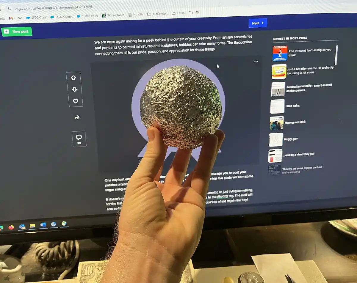 A hand holds a crumpled ball of aluminum foil directly in front of a computer monitor displaying an Imgur webpage promoting…