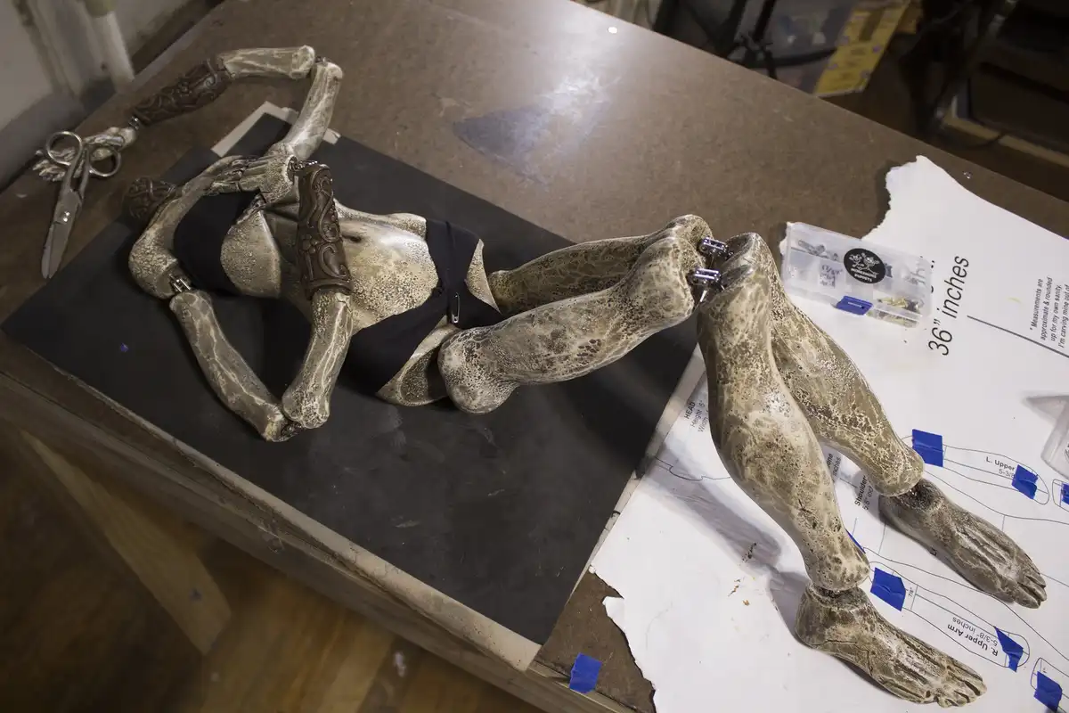 A partially carved, skeletal puppet figure lies on a workbench, surrounded by tools and blue-taped measurement papers. The…