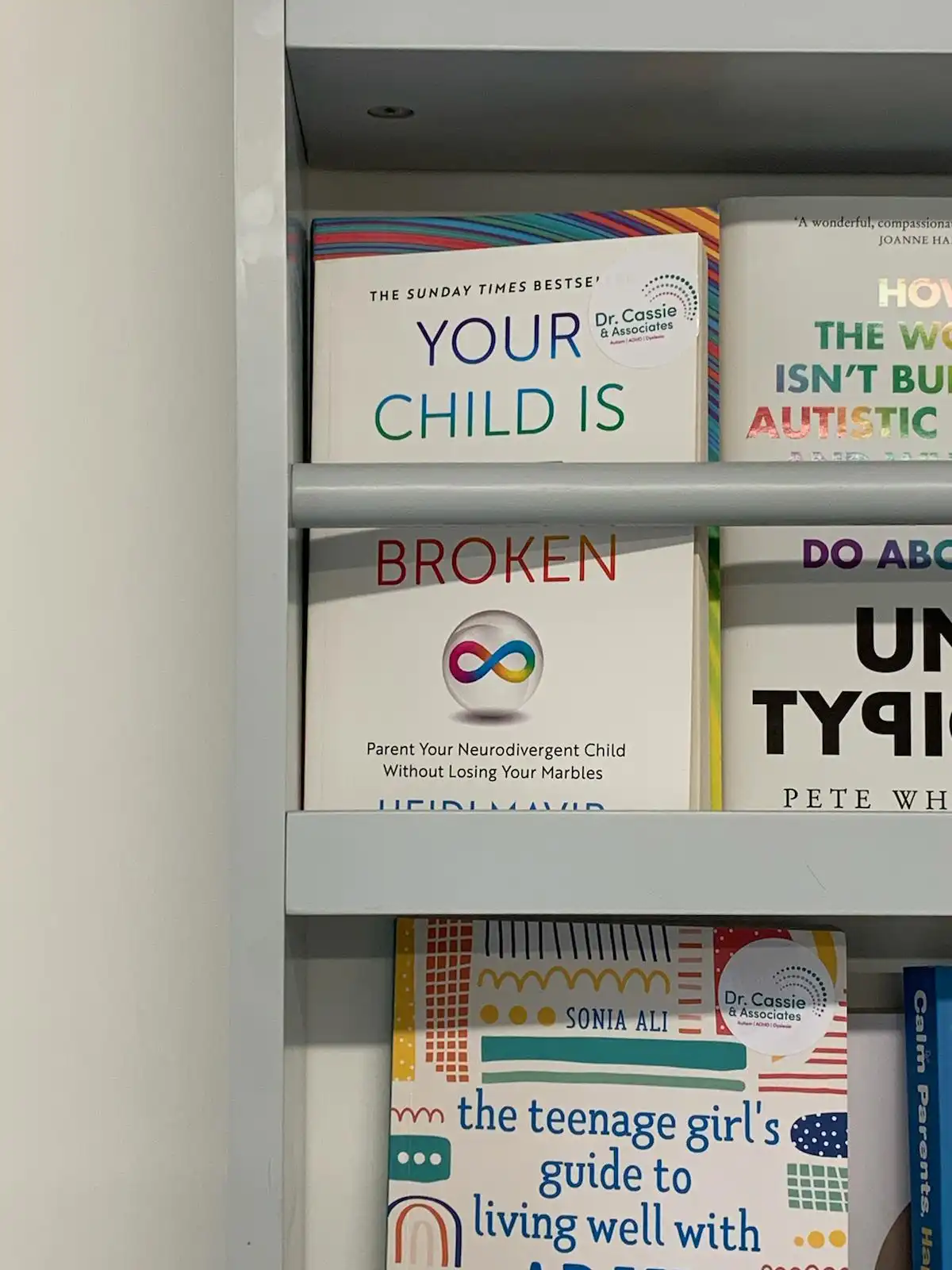 A bookshelf displays several parenting books about neurodivergent children, including one titled 'YOUR CHILD IS BROKEN' with…