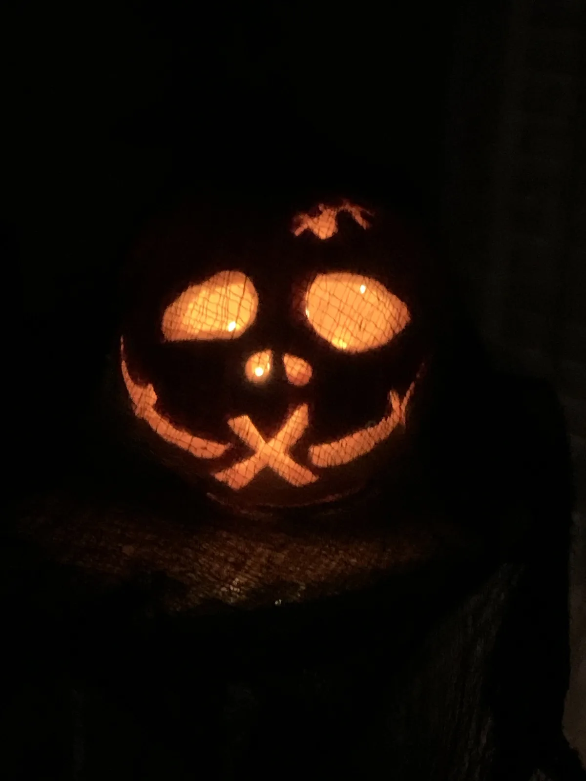 A carved pumpkin, illuminated from within by a warm light, displays a face with large oval eyes, a small nose, and a wide…