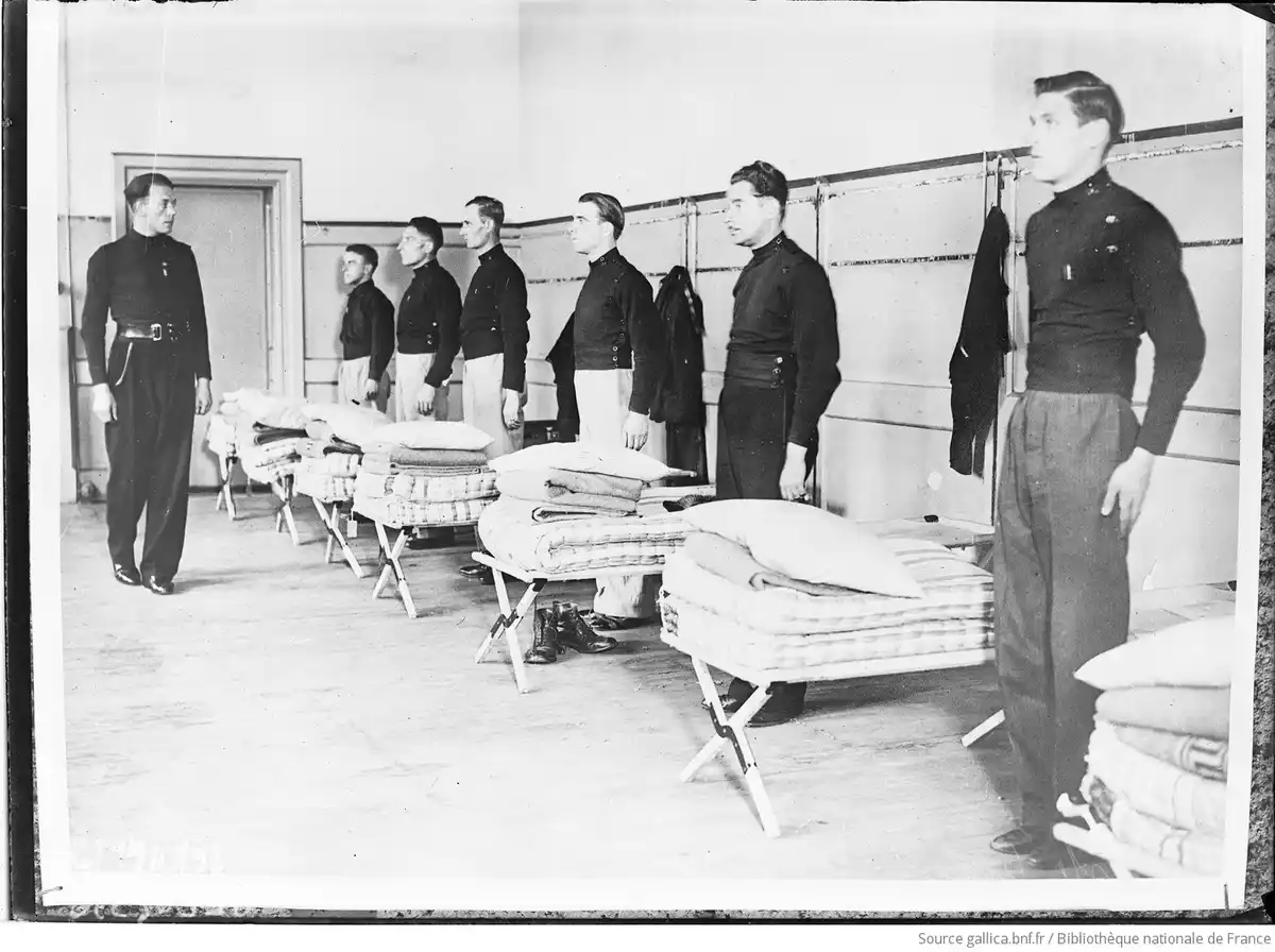 A black-and-white historical photo shows a line of uniformed men standing at attention in a barracks room with folding cots…