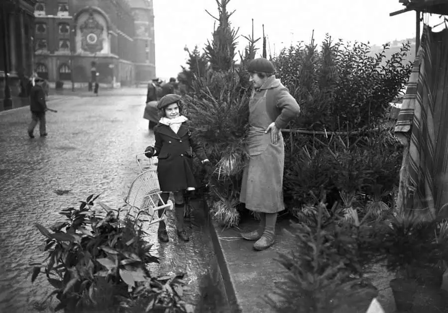 A vintage black-and-white photo shows a young girl in a coat and hat standing next to a small Christmas tree, smiling at the…