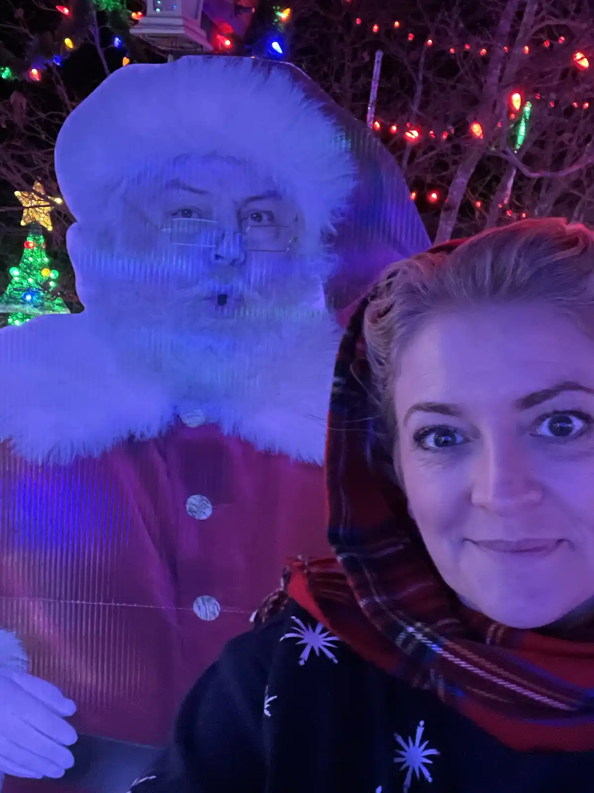 A woman takes a selfie at night in front of a large, illuminated Santa Claus cutout. The cutout has a man's face projected…