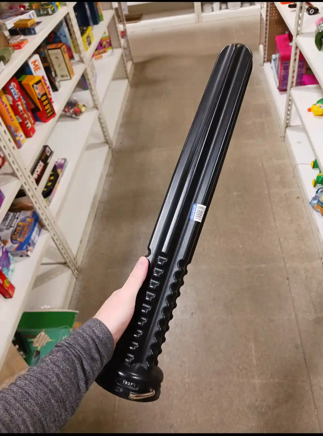 A person's hand holds a long, black, plastic bat with a textured grip and a barcode sticker, inside a store aisle lined with…