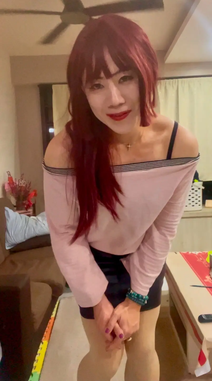 A woman with red hair and red lipstick is bending forward slightly in a living room, wearing a pink off-the-shoulder top and…