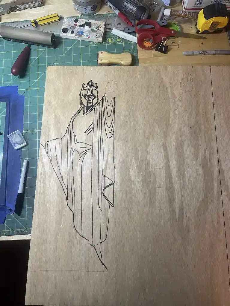 A top-down photo shows a wooden board with a black outline drawing of a stylized, robed figure resembling a villain or…
