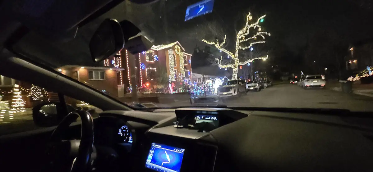 A nighttime dashcam view from inside a car driving down a residential street decorated with bright Christmas lights on…