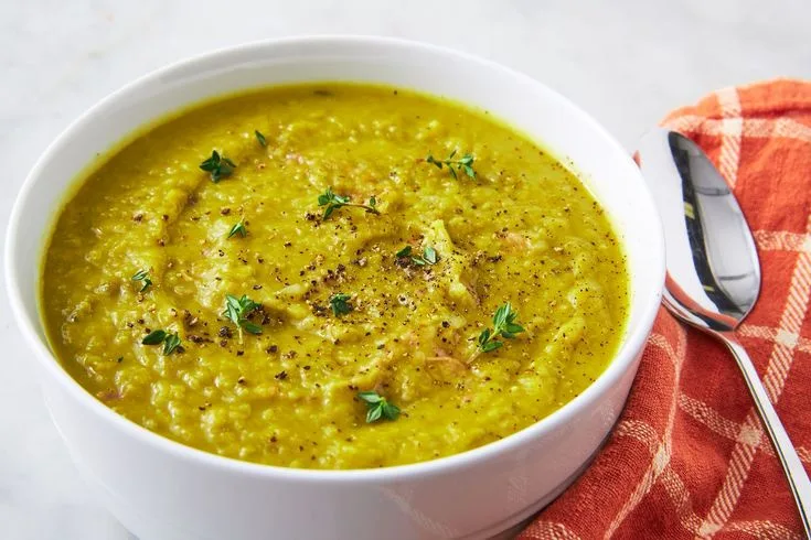 A white bowl filled with thick, yellow-green soup, garnished with fresh herbs and black pepper, sits on a white surface next…
