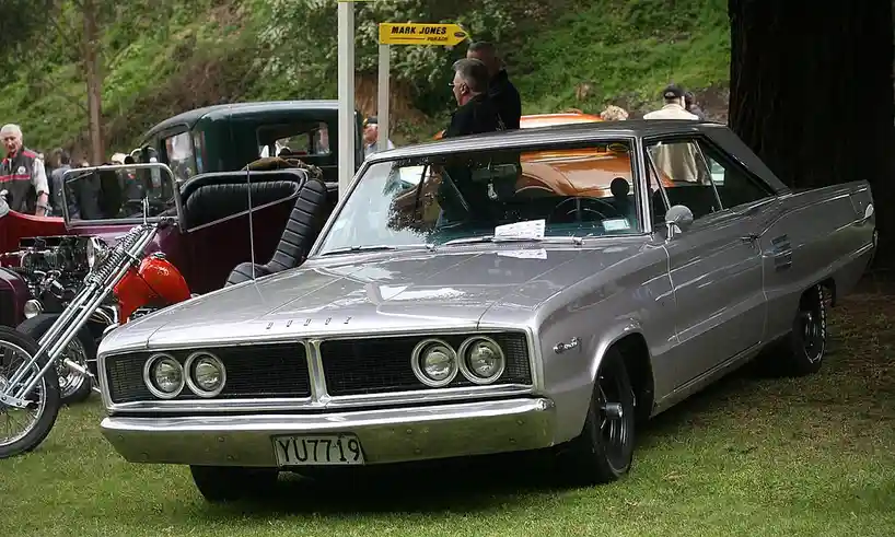 A silver 1960s Dodge automobile is parked on grass at a car show, with a yellow directional sign reading 'MARK JONES PARADE'…