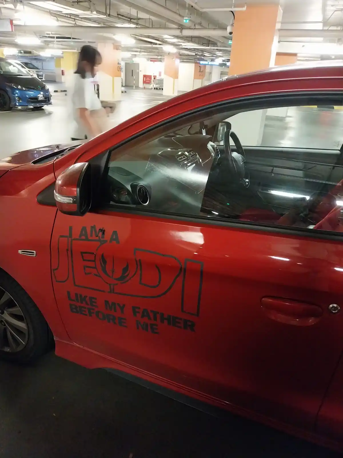 A red car parked in an underground garage has black text on its side door that reads 'I AM A JEDI LIKE MY FATHER BEFORE ME'…