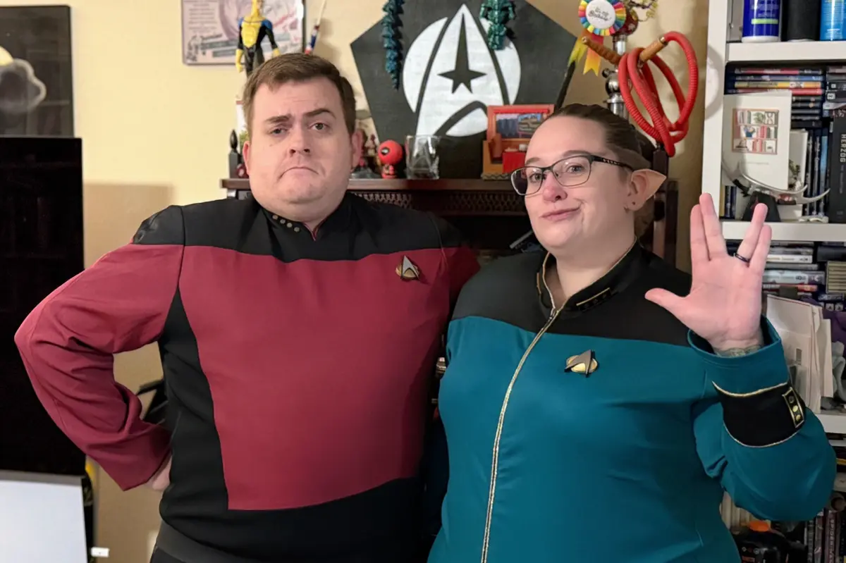 Two people dressed in Star Trek uniforms stand in a room decorated with Star Trek memorabilia. The person on the left wears…