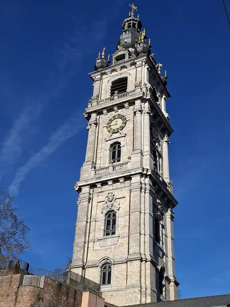 A tall, ornate stone clock tower with a visible clock face and golden accents stands against a clear blue sky. The overlaid…