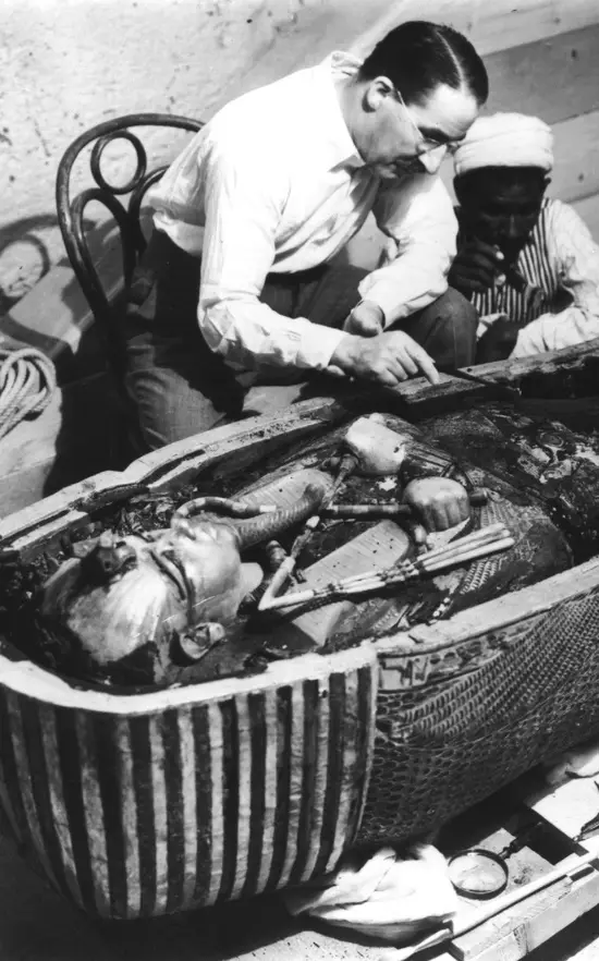 A black-and-white historical photo shows an archaeologist in a white shirt and glasses leaning over an open ancient Egyptian…
