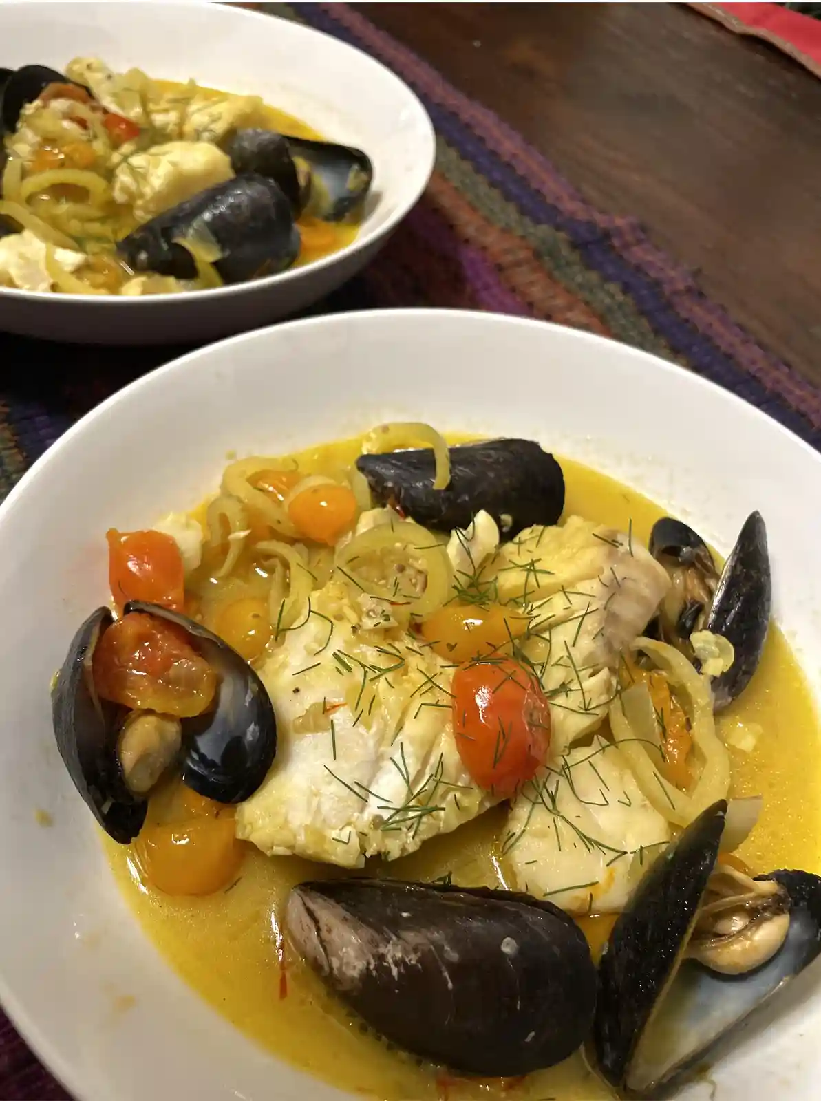 Two white bowls contain a seafood stew with mussels, chunks of white fish, cherry tomatoes, sliced onions, and fresh dill in…