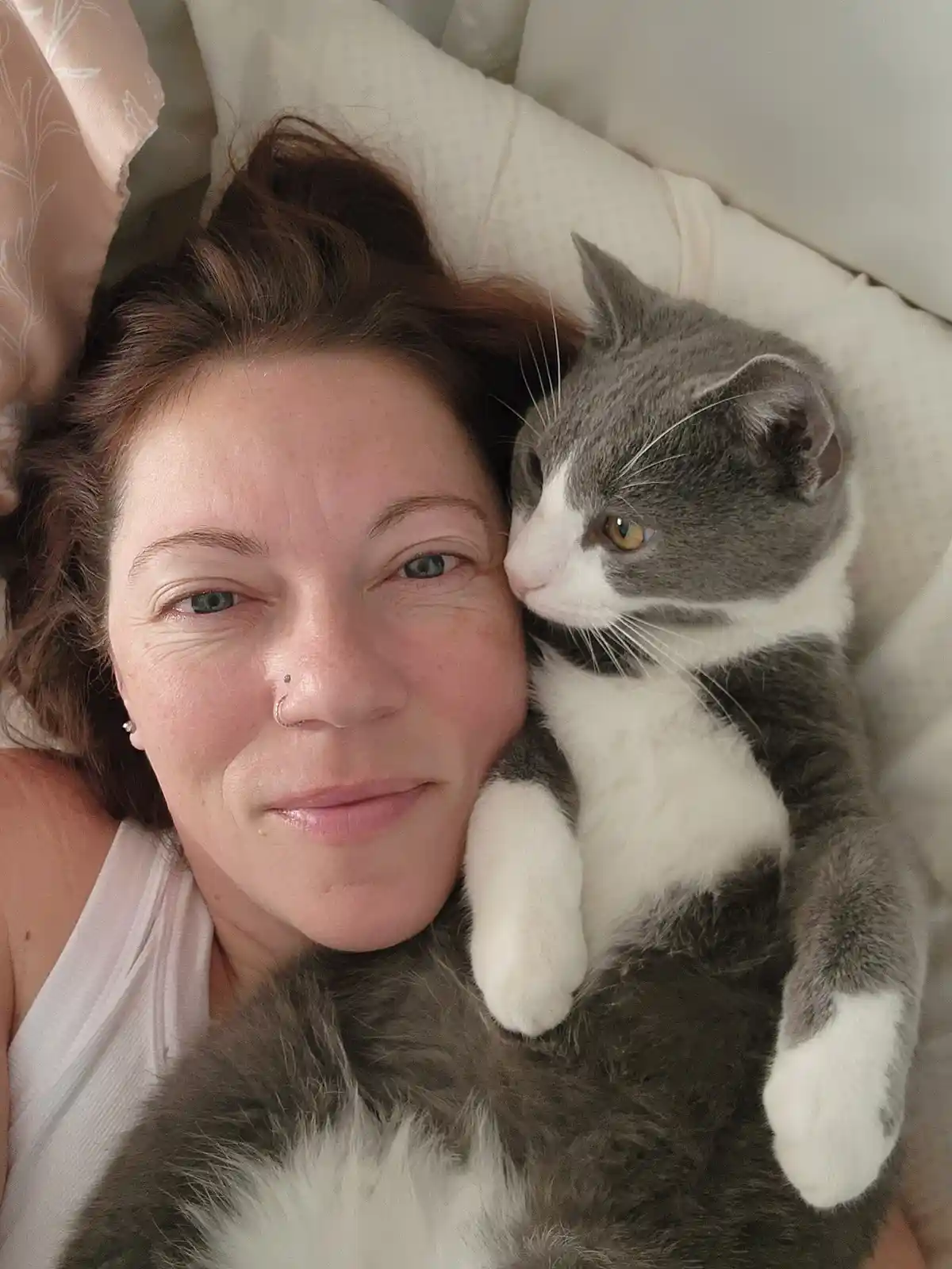 A woman with brown hair and a nose ring lies in bed, smiling at the camera while a gray and white cat rests its head on her…