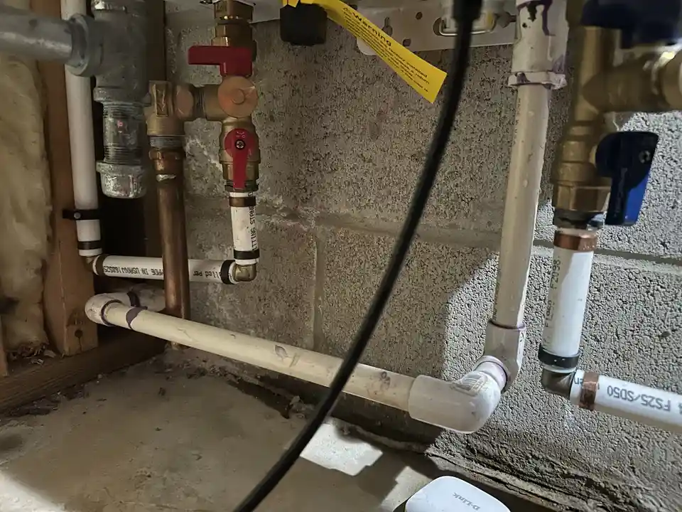 A close-up photo of a complex plumbing setup with copper and white PVC pipes, brass valves with red and blue handles, and a…