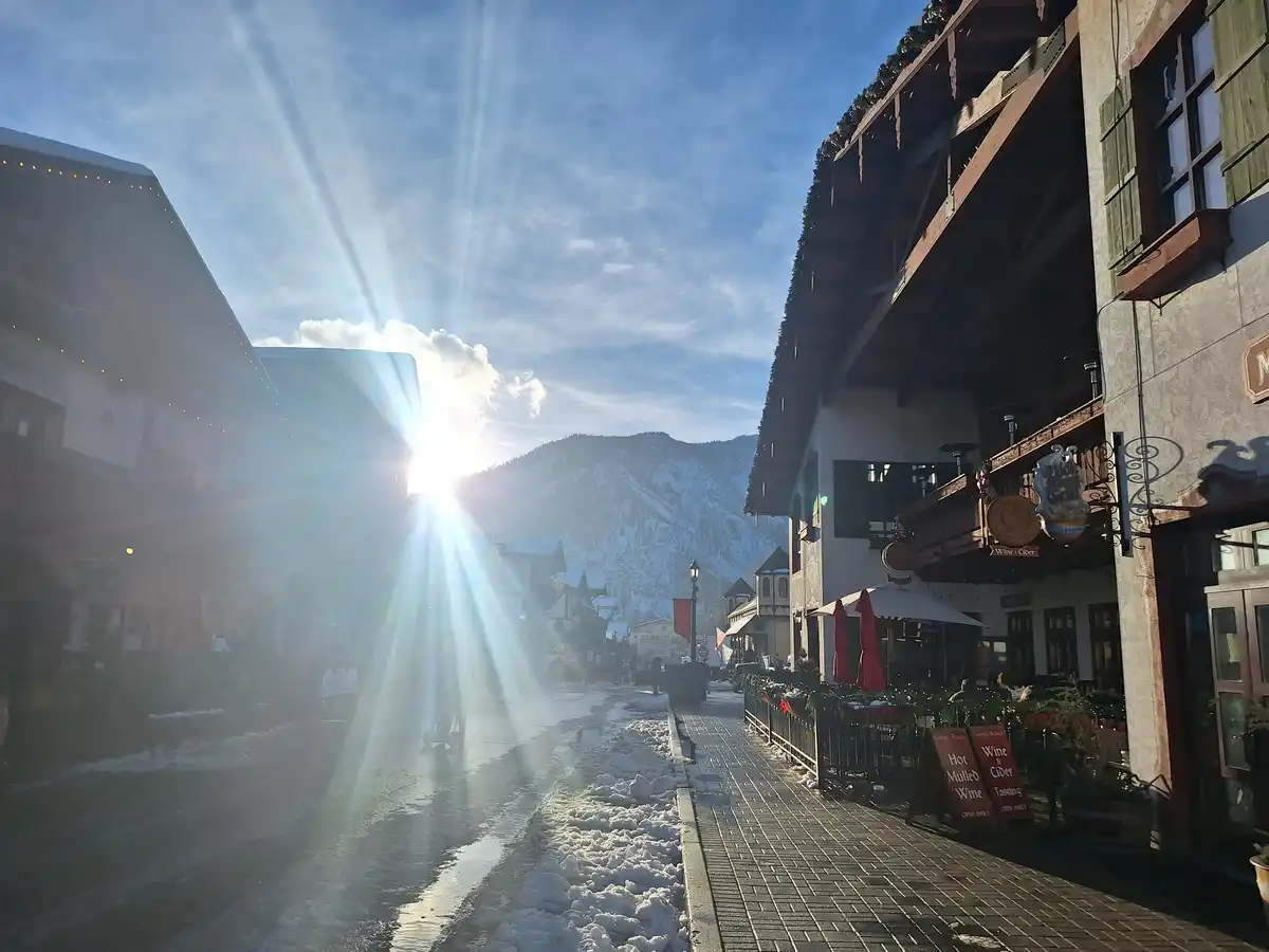 A bright sun shines down on a snowy, cobblestoned street lined with alpine-style buildings and outdoor seating, with…