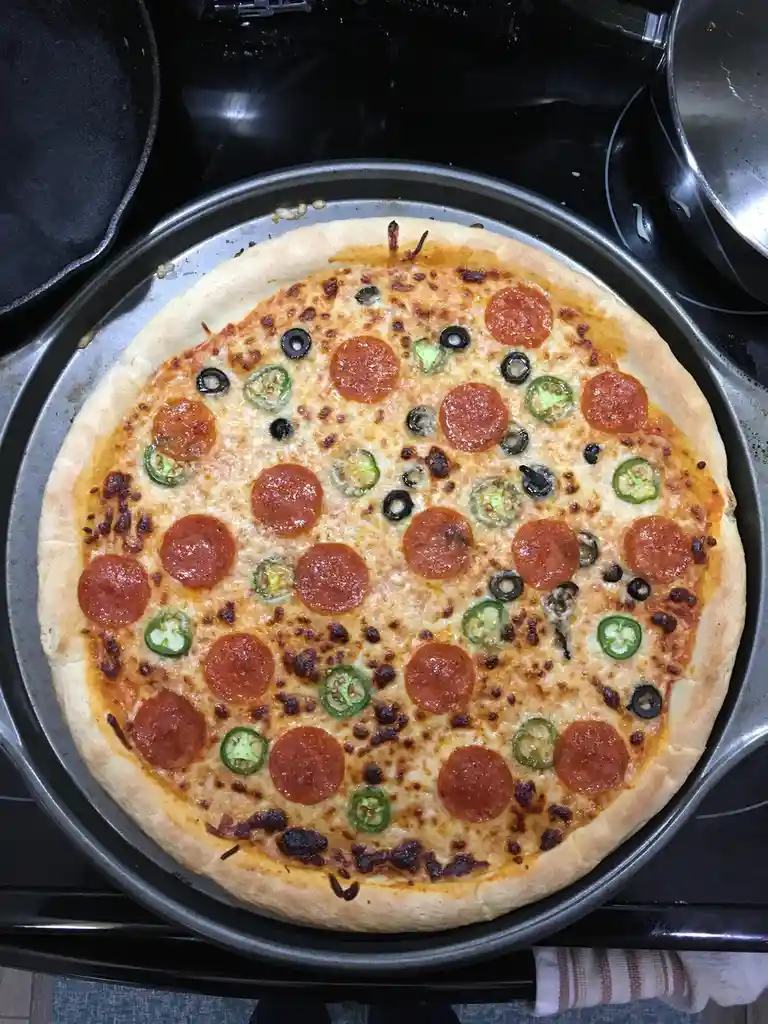 A freshly baked pepperoni, jalapeño, and black olive pizza sits on a metal baking pan atop a black stovetop. The cheese is…