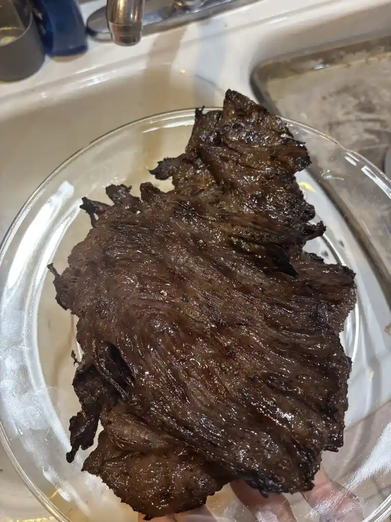 A hand holds a clear glass plate piled high with dark, charred, and visibly burnt strips of meat, likely beef, over a…