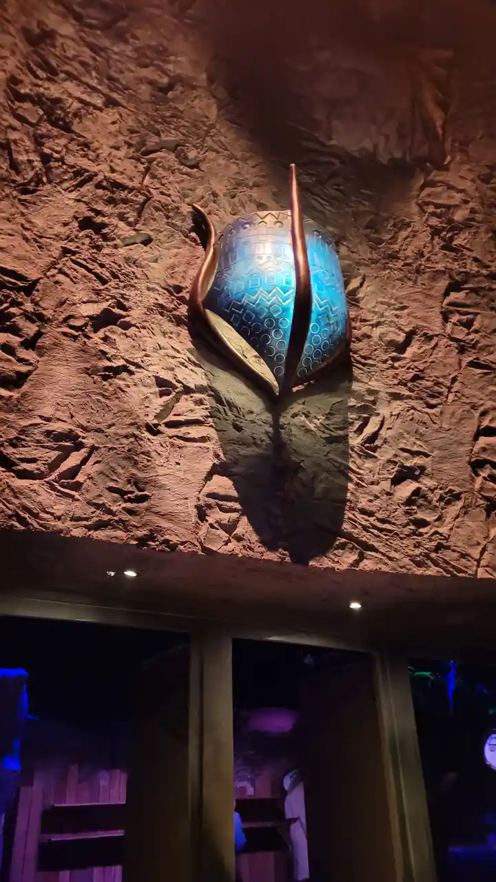 a large, glowing blue mask with intricate geometric patterns and bronze-colored horns is mounted on a textured…
