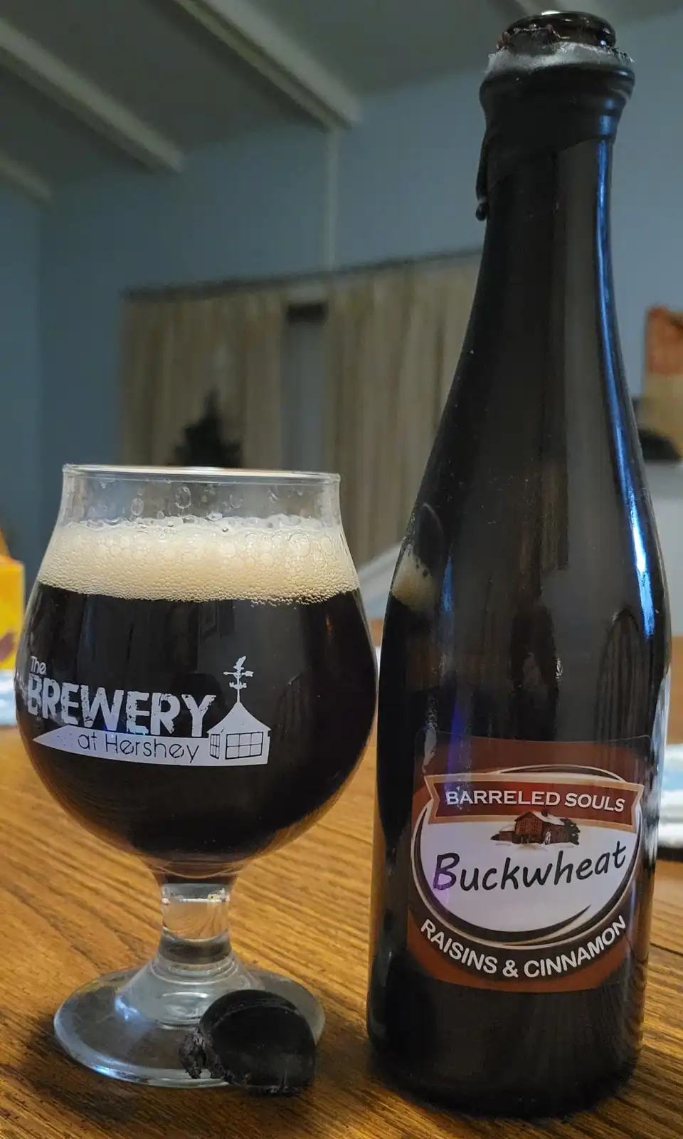 A dark beer bottle labeled 'Barreled Souls Buckwheat Raisins & Cinnamon' sits next to a glass from 'The Brewery at Hershey'…
