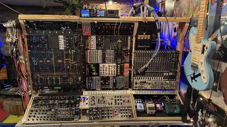 A densely packed wall of electronic music gear, including synthesizers, mixers, and effects pedals, with a light blue…