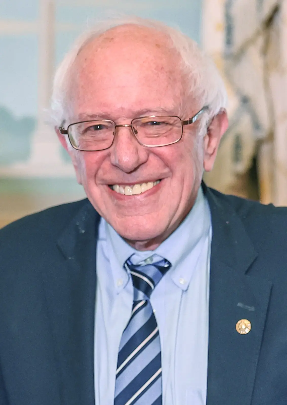 A smiling photo of Bernie Sanders wearing glasses, a blue suit, light blue shirt, and a striped tie. He has white hair and…