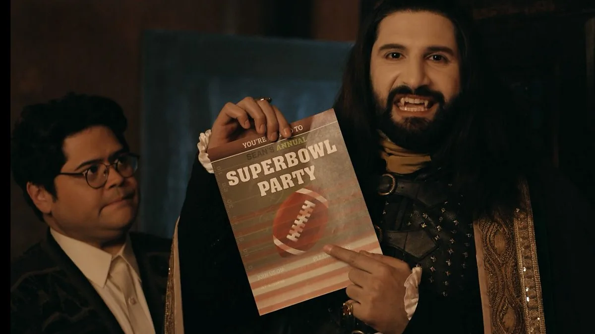 A man with long dark hair and a beard, dressed in a dark ornate costume, holds up a 'Super Bowl Party' invitation while…