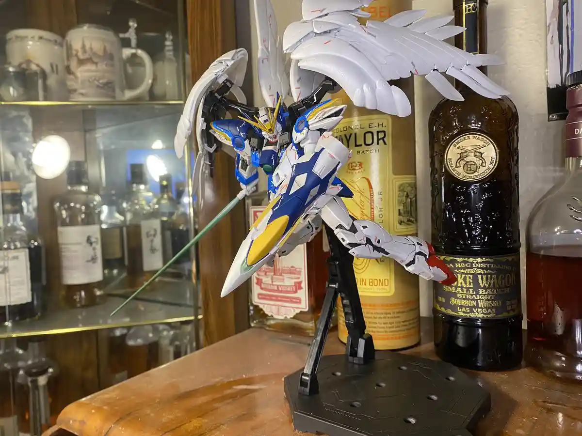 A blue, white, and yellow Gundam model with large white wing-like parts stands on a black display stand on a wooden surface…