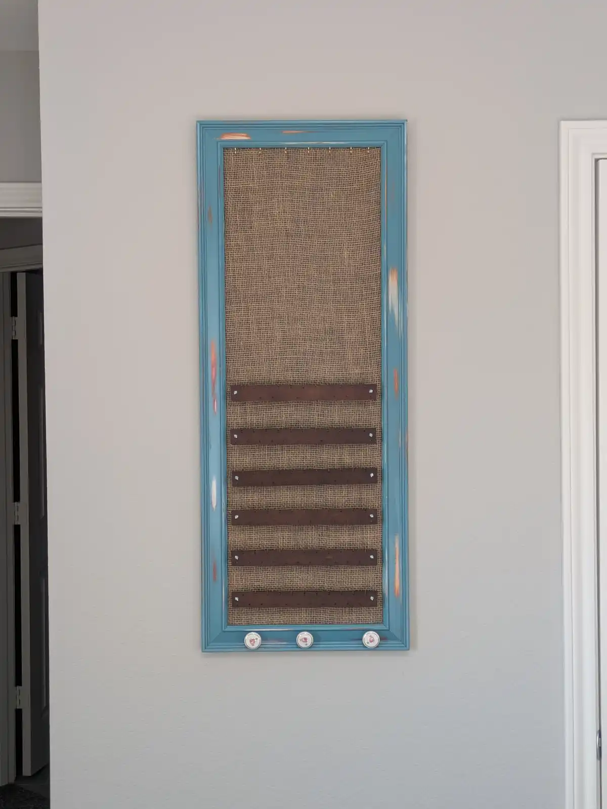 A tall, light blue wooden frame hangs on a white wall, containing a burlap-covered surface with five horizontal wooden…