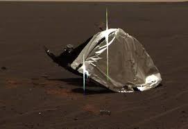 a crumpled, silver, reflective material lies on reddish-brown Martian soil, with two thin green poles stuck into the ground…