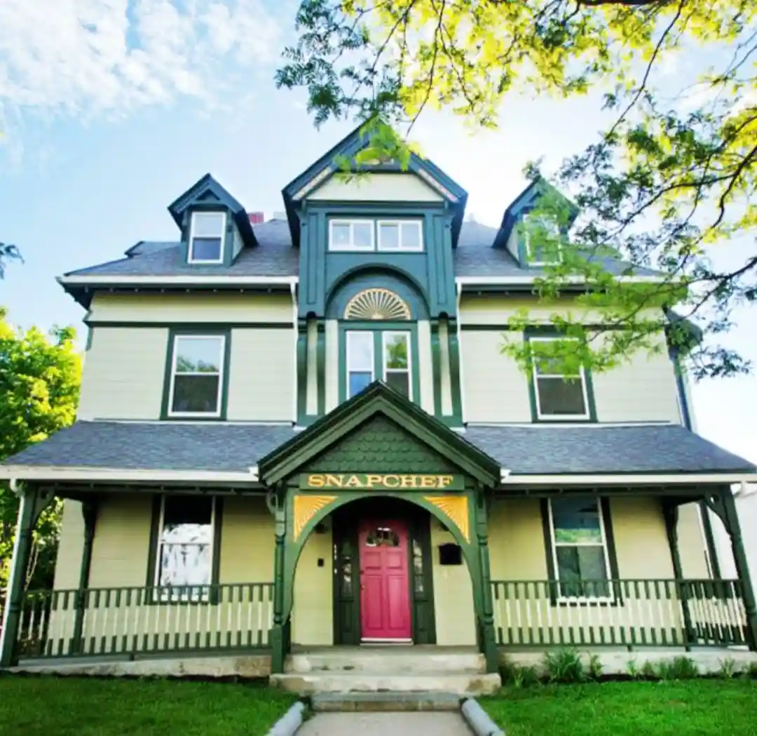 A large, two-story Victorian-style house with light green siding and dark green trim, featuring a prominent porch with a…