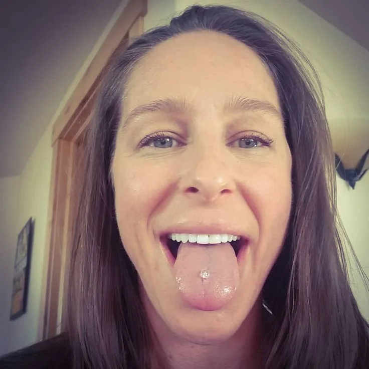A close-up selfie of a woman with long brown hair, smiling broadly while sticking her tongue out to show a small silver…