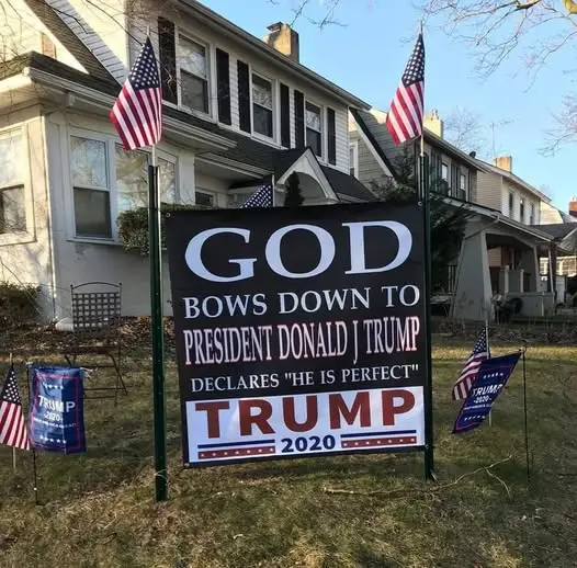 A large black yard sign with white and red text stands in a grassy front yard, flanked by American flags and smaller Trump…