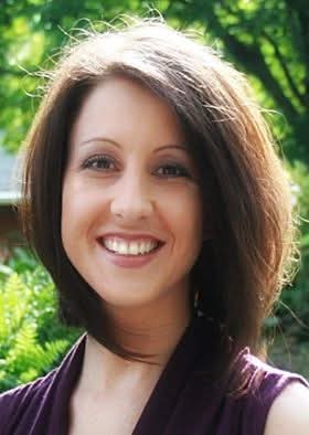 A smiling woman with shoulder-length brown hair wearing a purple top, standing outdoors with green foliage in the…