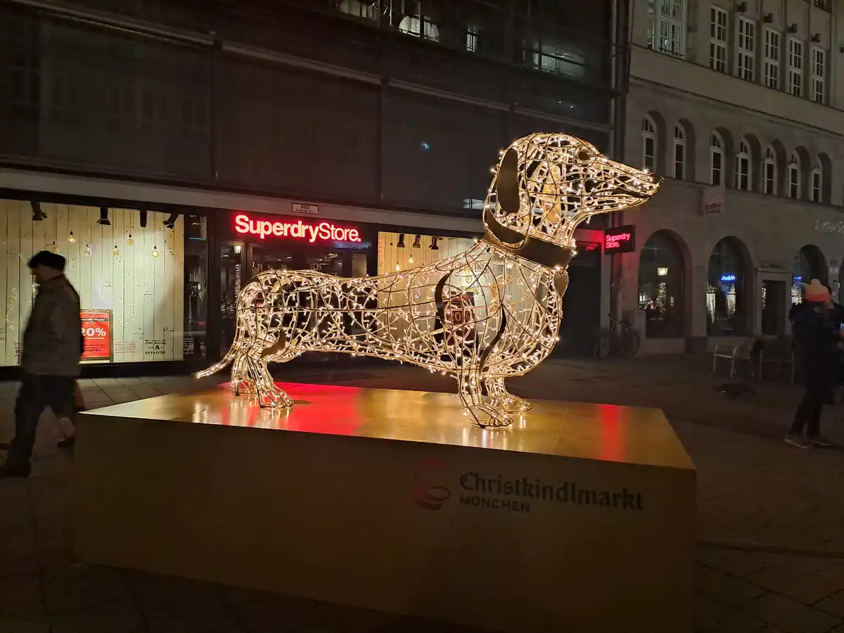 A large, illuminated wireframe sculpture of a dachshund stands on a pedestal labeled 'Christkindlmarkt MÜNCHEN' at night…