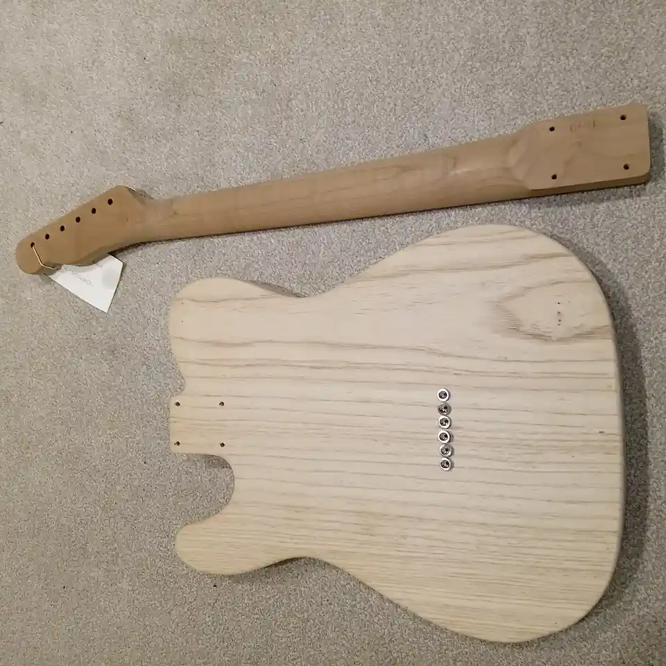 A top-down photo shows a bare, unfinished electric guitar body and neck lying on a gray carpet. The body has a light wood…