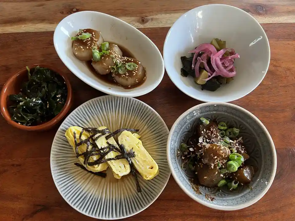Five small dishes of Japanese food are arranged on a wooden table: a bowl of simmered scallops with sesame seeds and green…