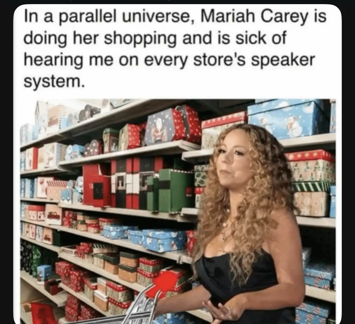 A woman with long curly hair, identified as Mariah Carey, stands in a store aisle filled with colorful Christmas gift boxes…