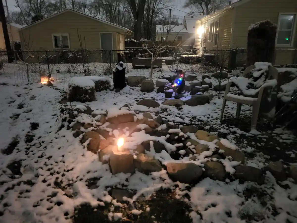 A snowy backyard at night features a circle of rocks with a single lit candle in the center, surrounded by snow. In the…