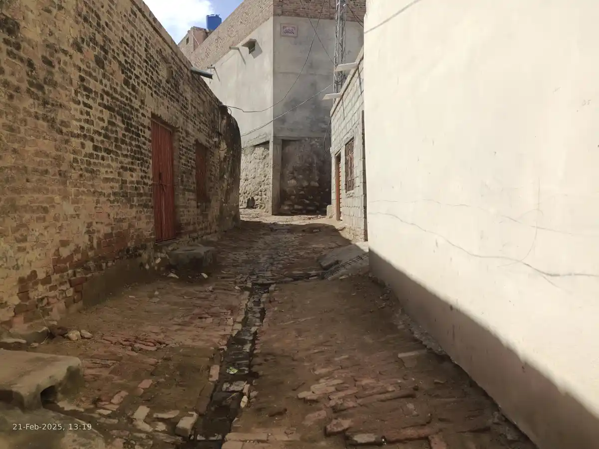 A narrow, sunlit alleyway with uneven brick pavement, flanked by weathered brick and white walls, showing a small puddle in…