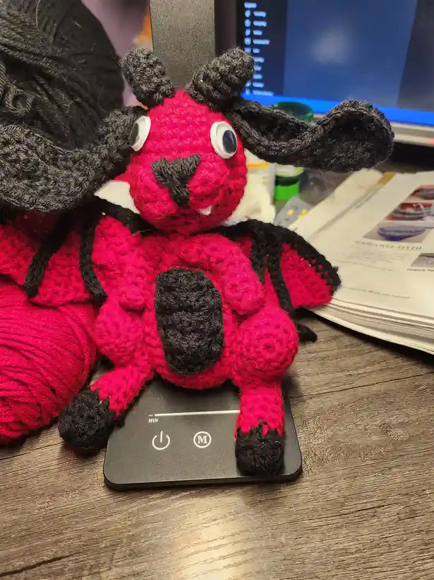 A handmade, crocheted gargoyle plush toy, primarily red with black wings, horns, and feet, sits on a small black electronic…