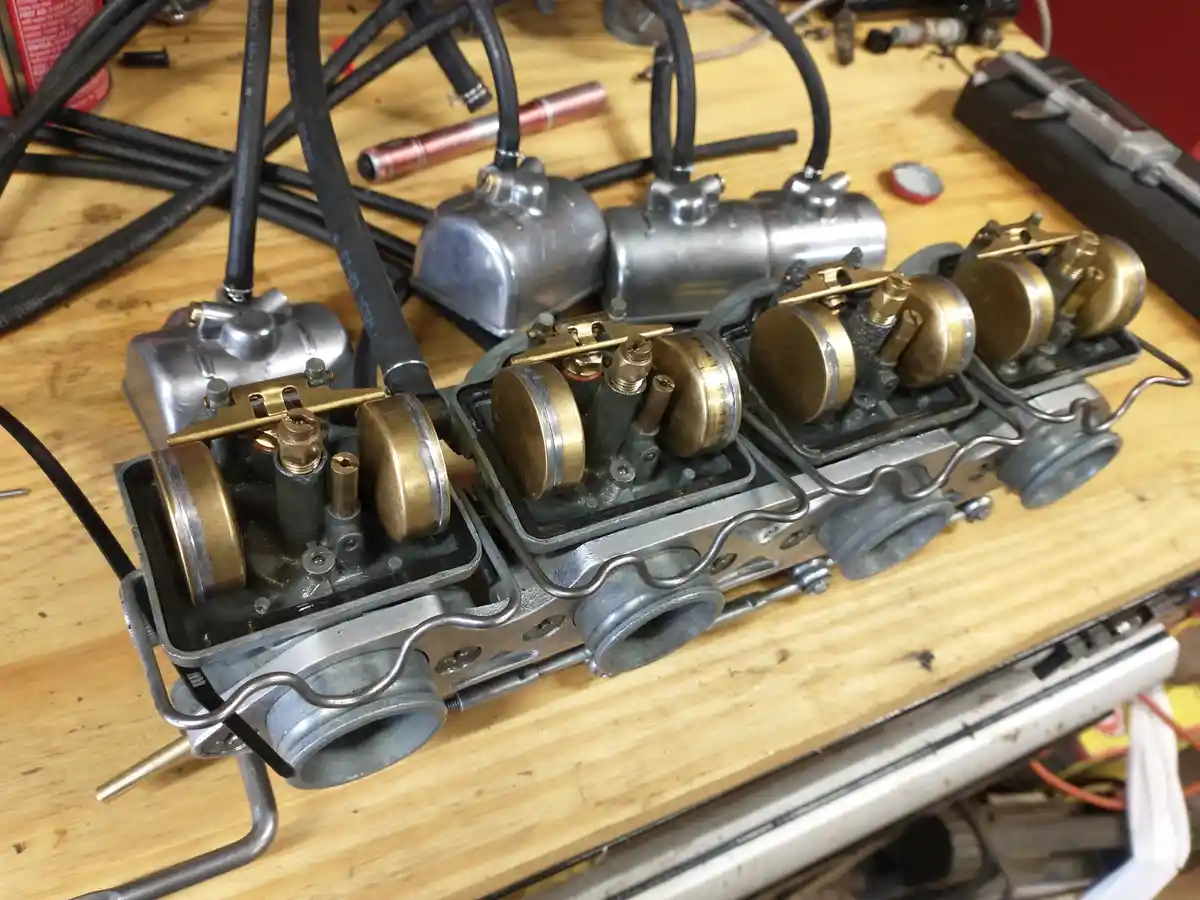 A close-up photo shows a disassembled and partially reassembled engine manifold on a wooden workbench, surrounded by tools…