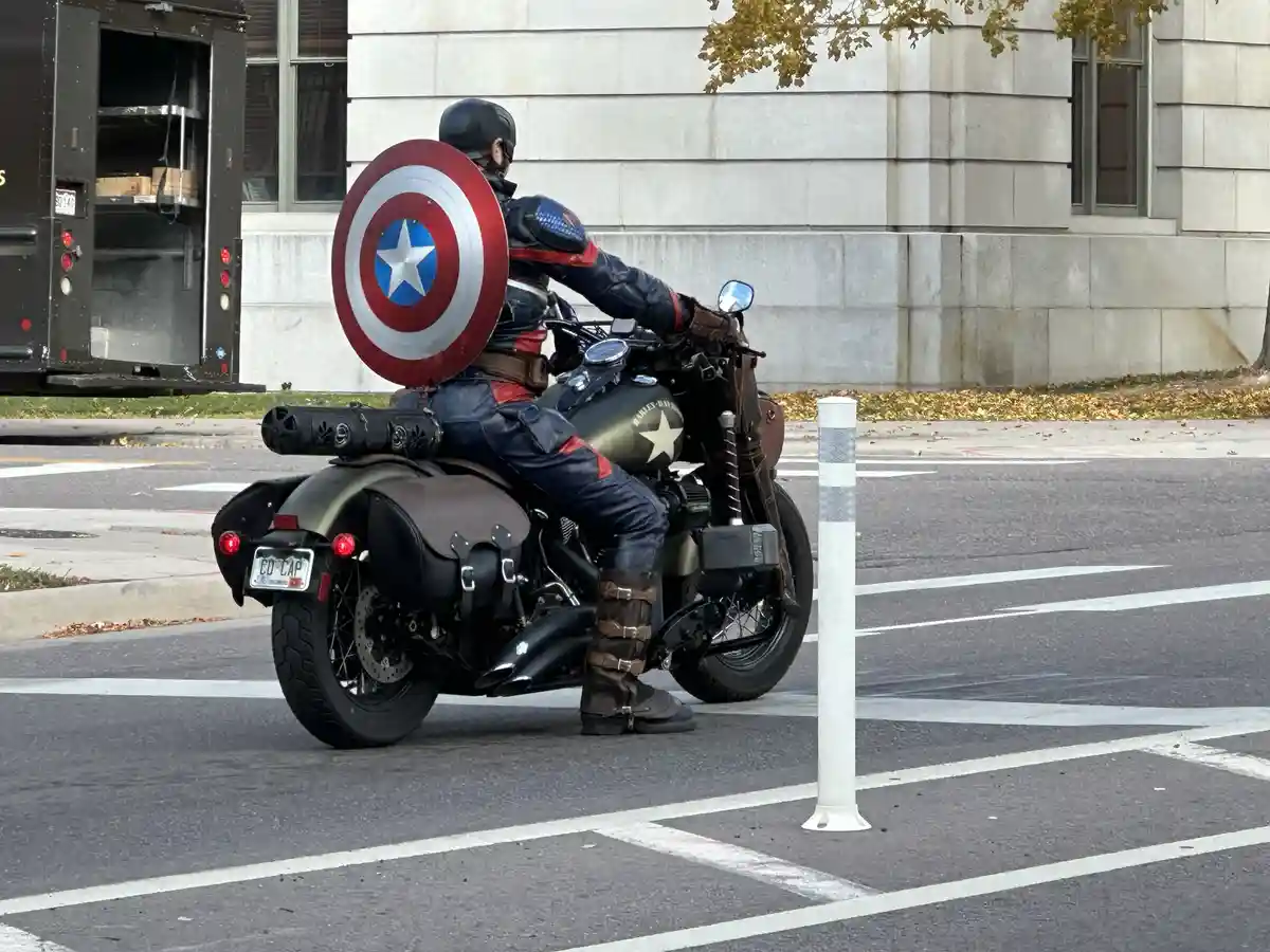 A person dressed as Captain America, wearing his iconic helmet and holding his circular shield, is seated on a customized…