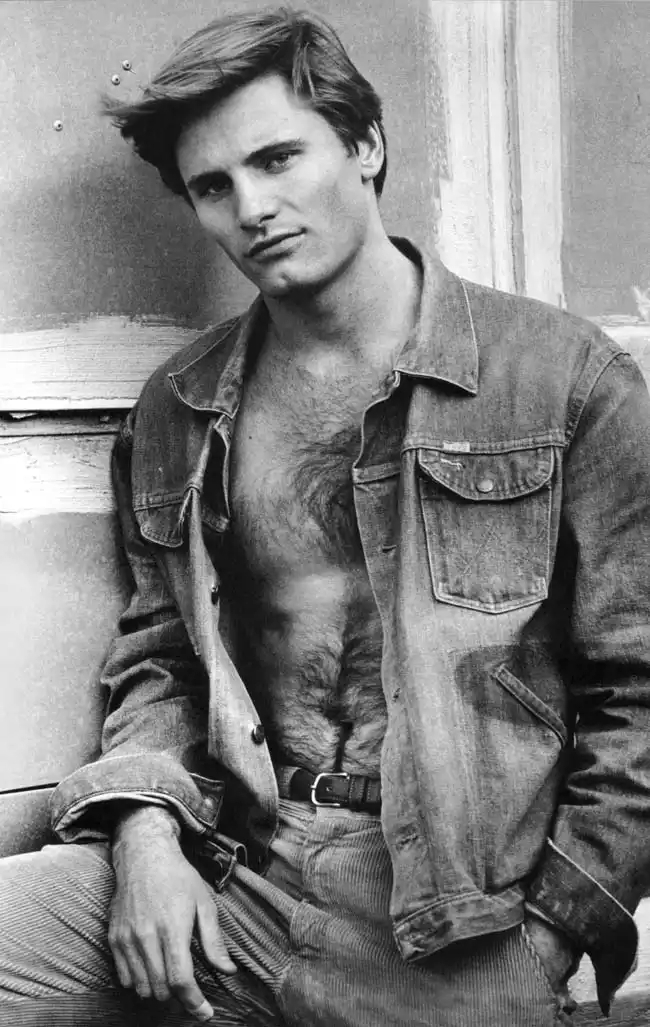 A black-and-white photo of a young man with a serious expression, wearing an unbuttoned denim jacket revealing his hairy…