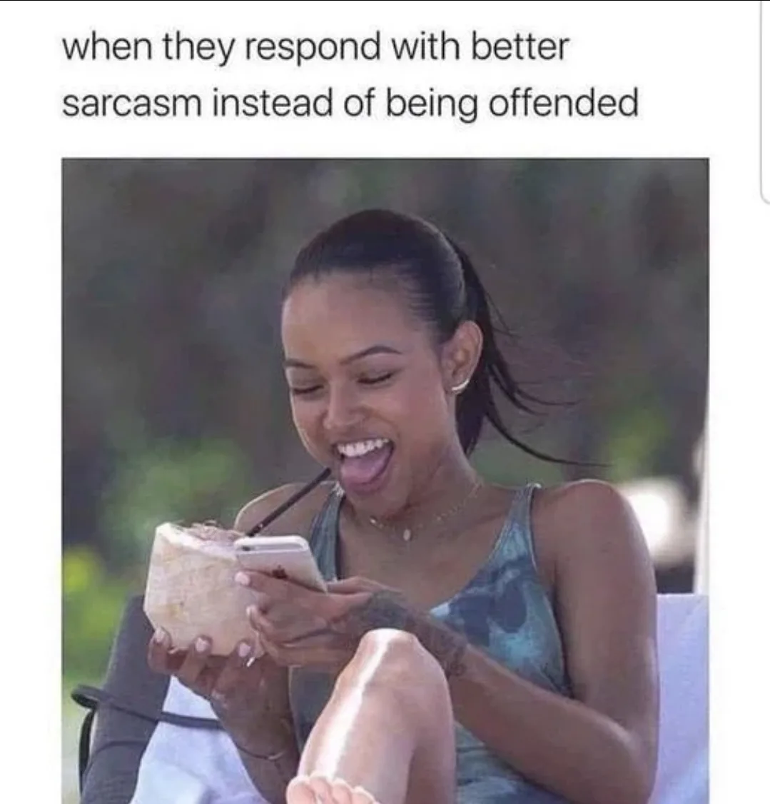 A woman with dark hair in a ponytail, wearing a green tank top, sits outdoors holding a coconut with a straw and a…