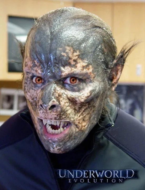 A person wearing a detailed, grotesque werewolf or vampire-like makeup with textured grey and brown skin, pointed ears, and…