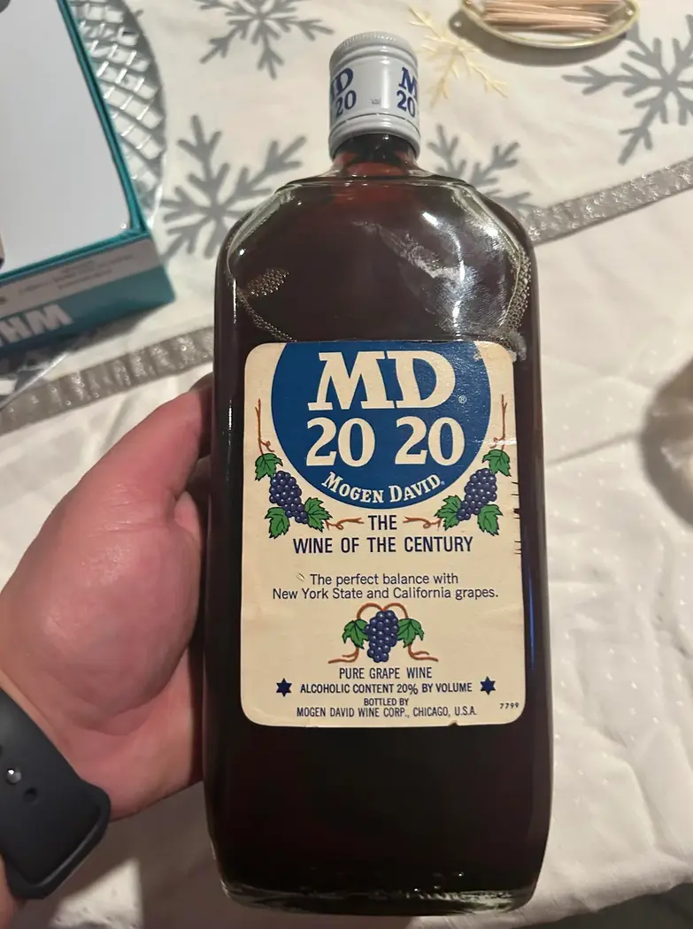A hand holds a bottle of Mogen David 'MD 20 20' wine, labeled 'The Wine of the Century' with 20% alcohol content, against a…